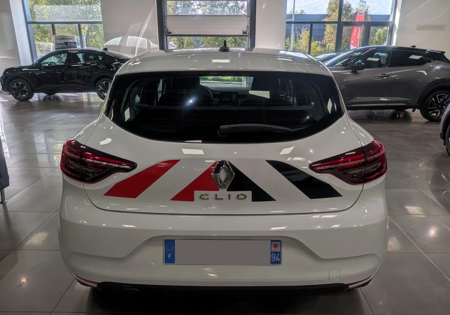 Vue arrière d'une Renault Clio V blanche Glacier avec marquages rouges et noirs distinctifs sur le coffre, en intérieur showroom.
