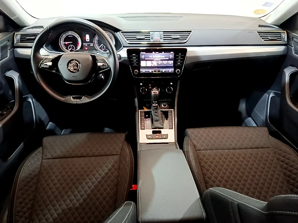 Intérieur de la Skoda Superb 2020 vu de face, tableau de bord noir avec écran tactile et sièges en tissu matelassé.