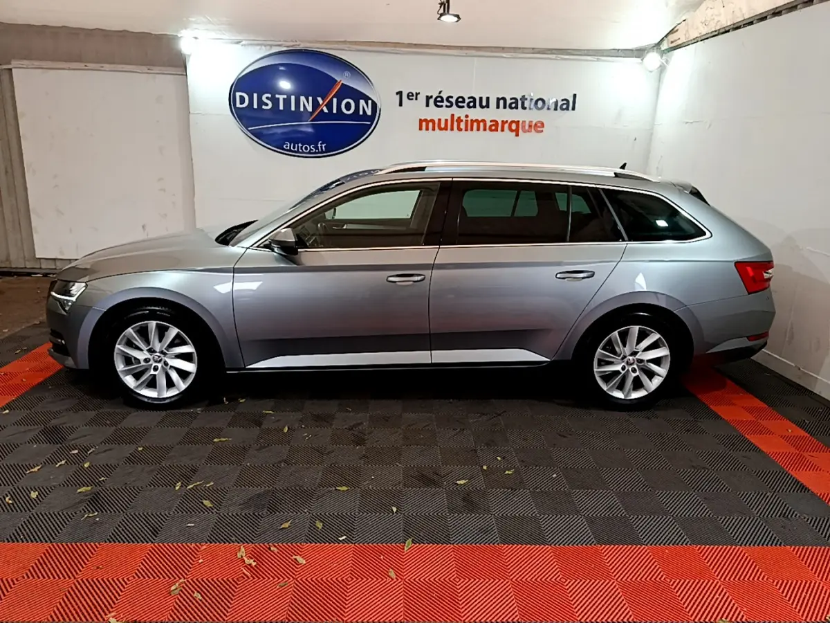 Profil latéral droit d'une Skoda Superb gris clair en version break, stationnée en intérieur sur sol noir et rouge.