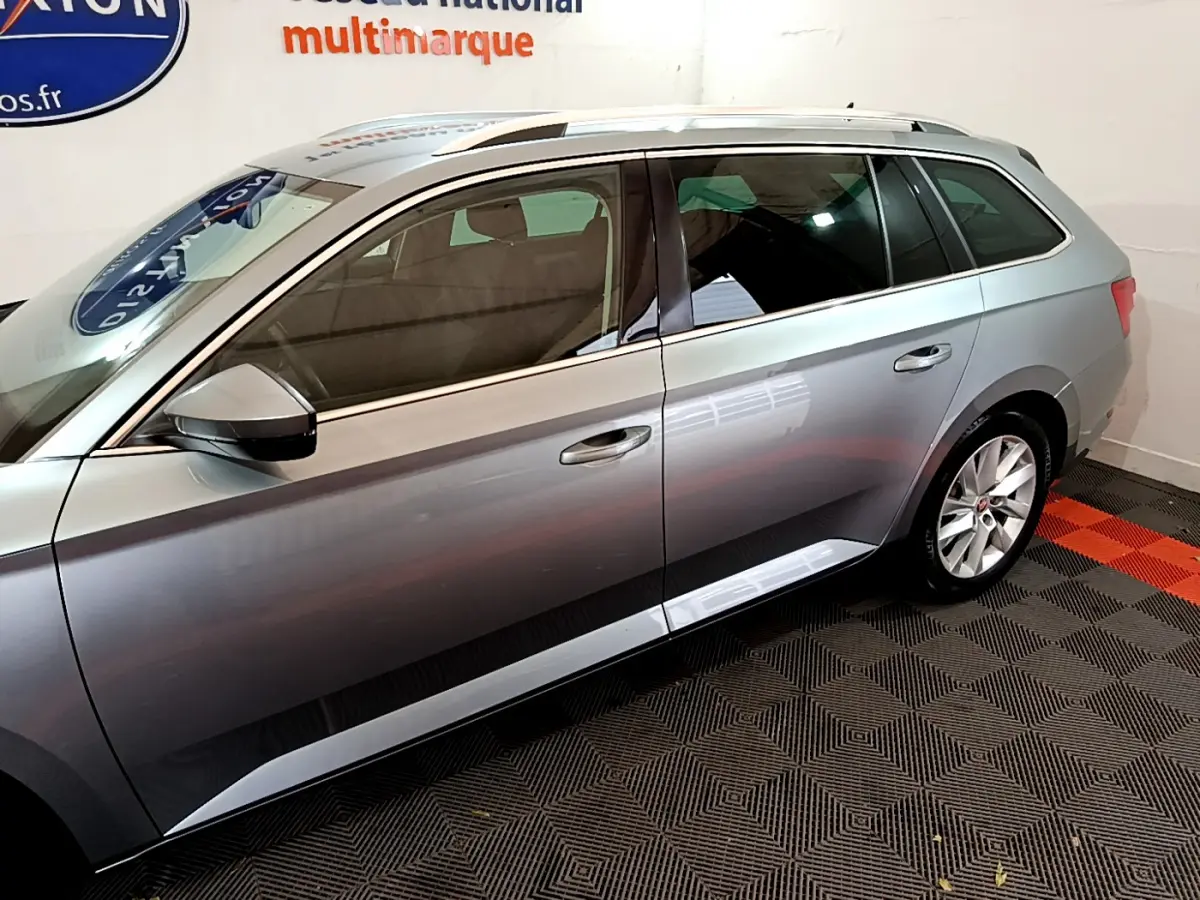 Profil côté gauche d'une Skoda Superb gris clair 2020 en stationnement intérieur sur sol damier noir et rouge.