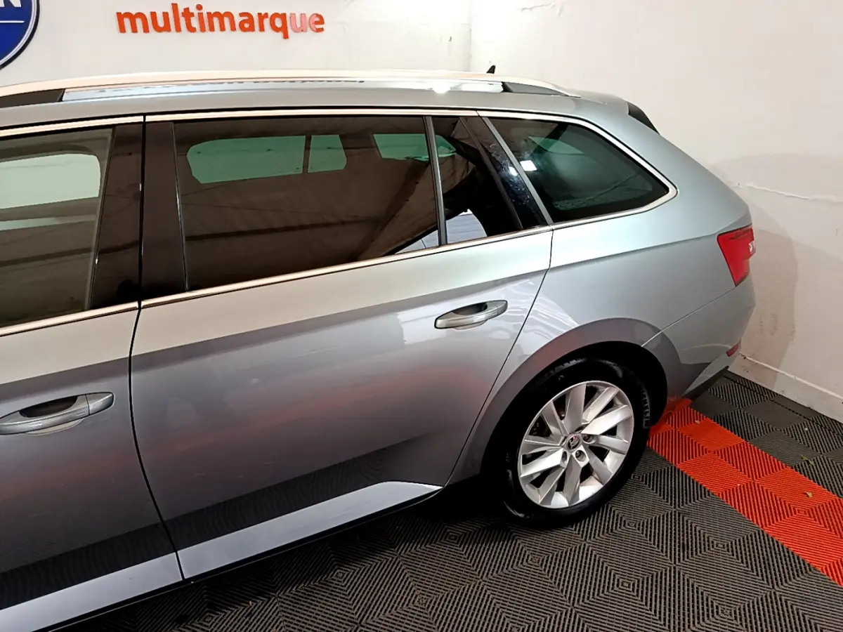 Profil côté gauche d'une Skoda Superb gris clair stationnée sur un sol à motifs noirs et rouges en intérieur.