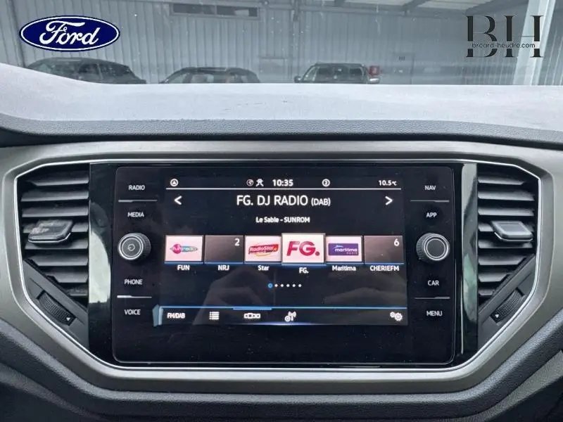 Tableau de bord avec écran tactile central allumé, affichant la radio FG DJ dans un Volkswagen T-Roc bleu Ravenna.