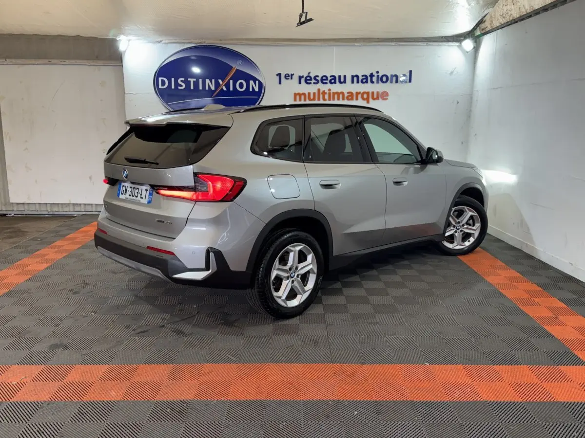 BMW X1 sDrive20i gris foncé vue 3/4 arrière droit dans un showroom avec éclairage mural et logo Distinxion.