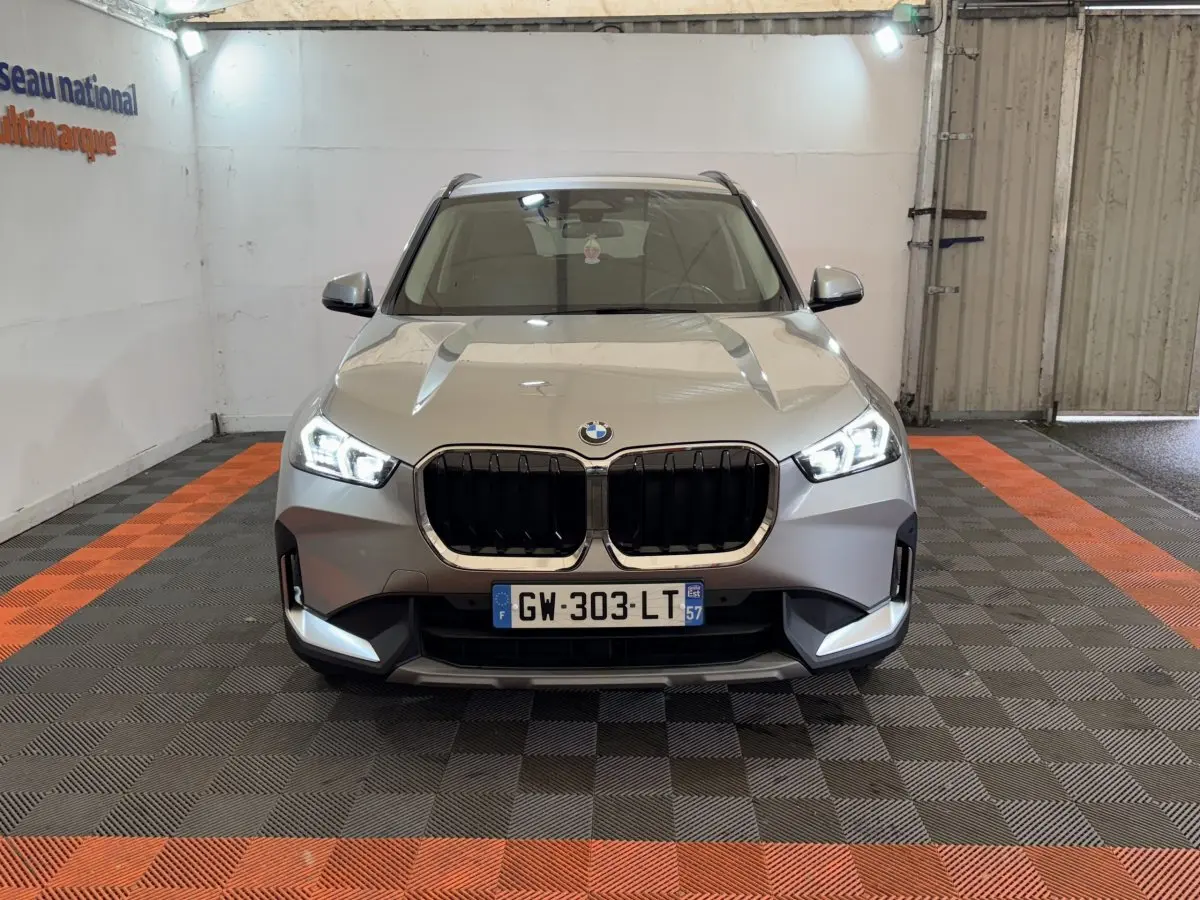 Vue frontale d’un BMW X1 gris foncé 2024 avec calandre noire et phares LED allumés dans un garage.