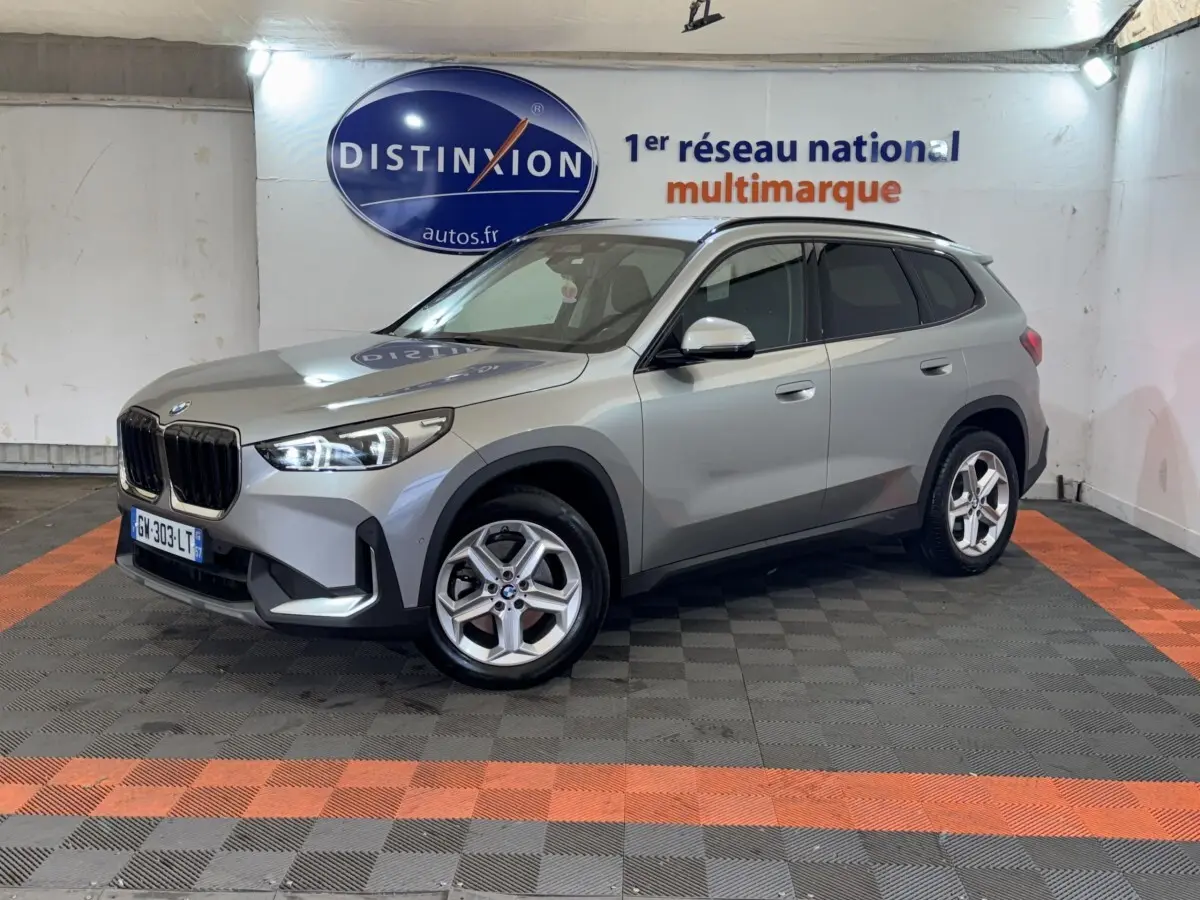 BMW X1 sDrive20i Business Design gris foncé vue 3/4 avant droit dans un showroom avec logo Distinxion en arrière-plan.