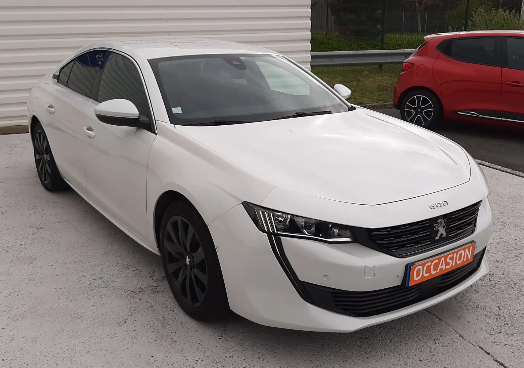 Peugeot 508 blanc vue 3/4 avant droit, calandre noire distinctive et jantes foncées, stationnée devant un mur blanc.