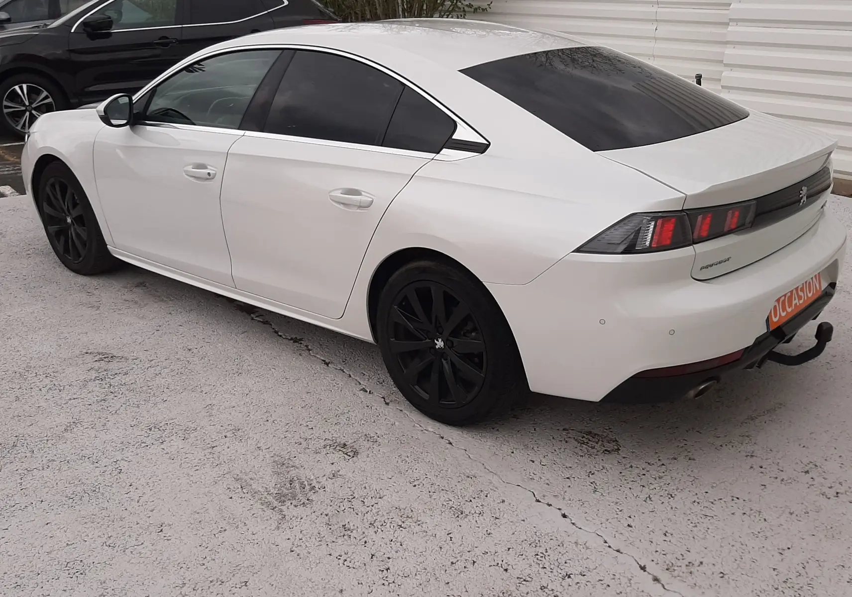Vue 3/4 arrière droite d'une Peugeot 508 blanche avec jantes noires et vitres teintées, stationnée sur sol clair.