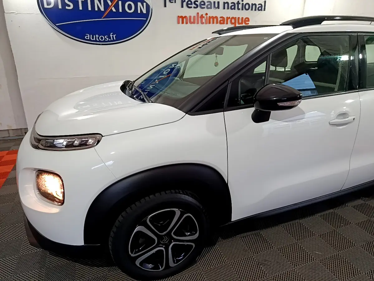 Vue de profil avant droit d'une Citroën C3 Aircross blanche avec feux avant allumés et rétroviseurs noirs.