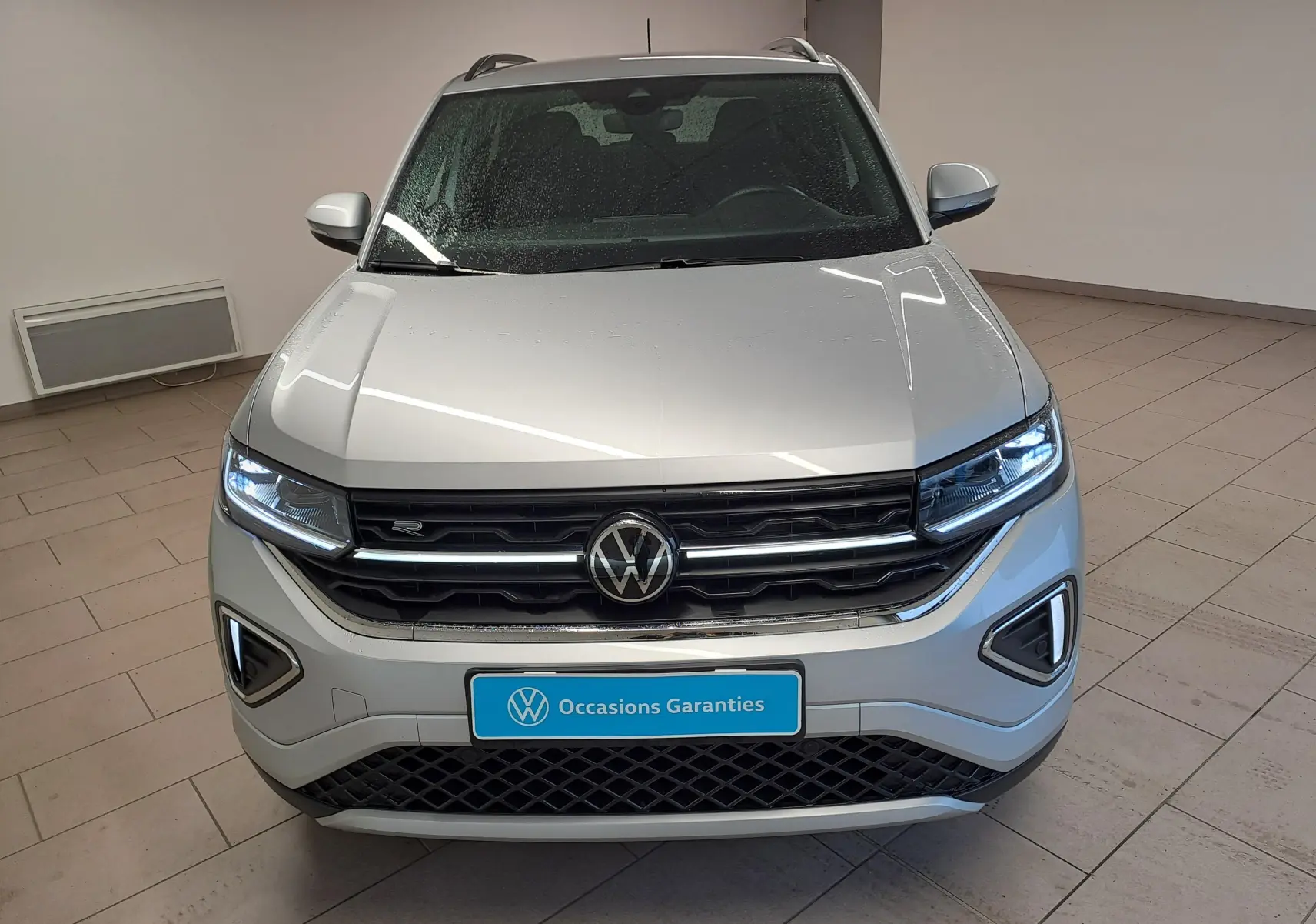 Vue avant d'un Volkswagen T-Cross 2025 R-Line Edition en Reflet d'argent avec phares allumés dans un showroom.