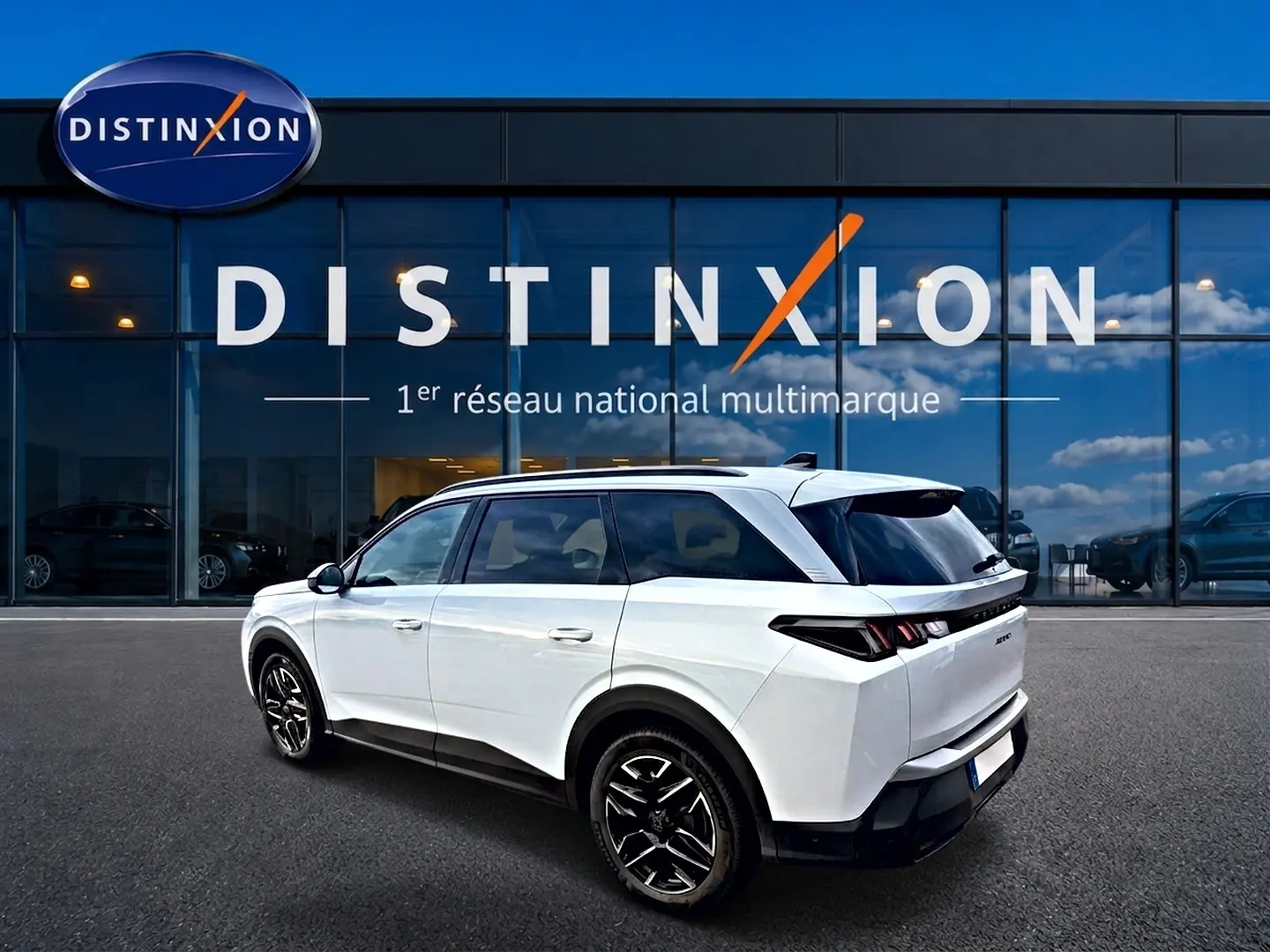 Peugeot 5008 Hybrid blanc en 3/4 arrière droit, avec jantes alu noires et toit noir devant une vitrine Distinxion.