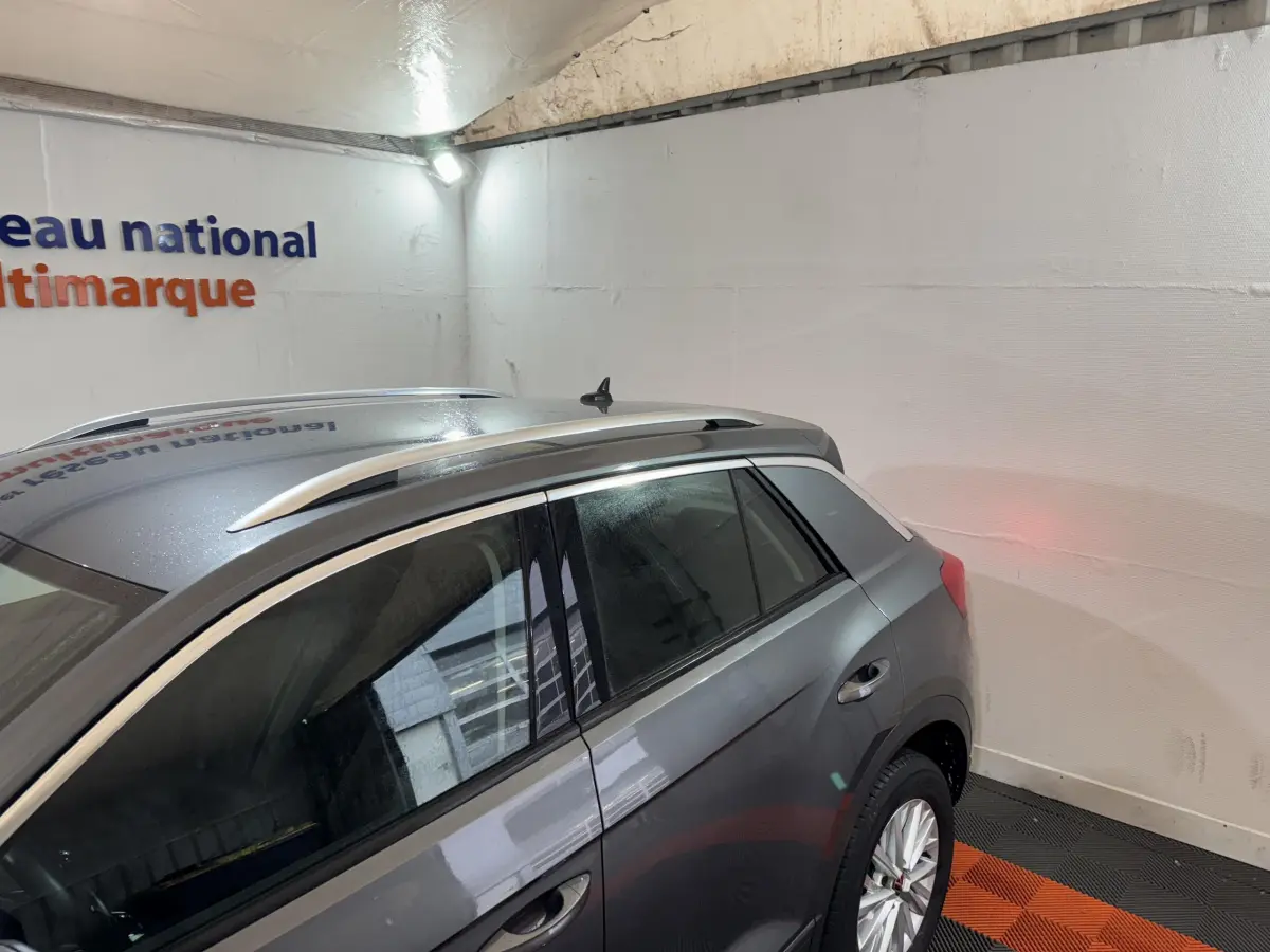 Vue 3/4 arrière droite d'un Volkswagen T-Roc gris foncé avec barres de toit argentées dans un garage.