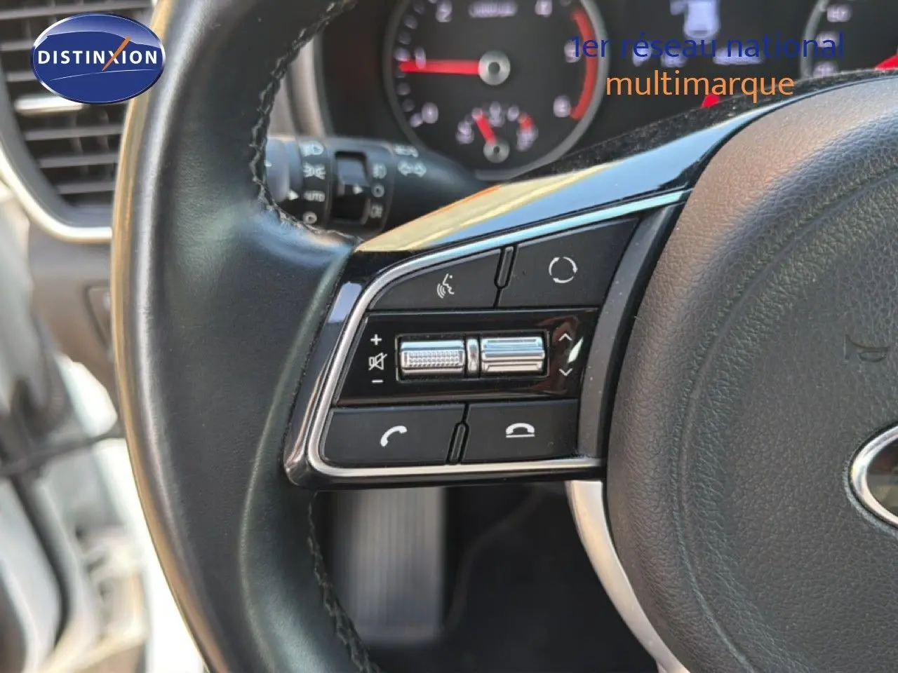 Gros plan sur les commandes gauche du volant noir du Kia Sportage blanc nacré, avec tableau de bord flou en arrière-plan.