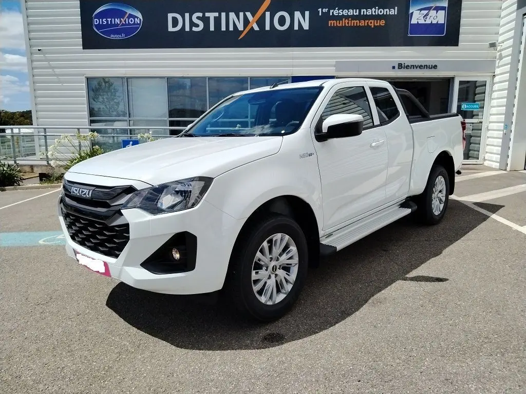 Vue de profil côté droit d'un pick-up Isuzu D-MAX blanc avec arceau noir et couvre-benne roulant, stationné devant un bâtiment industriel.