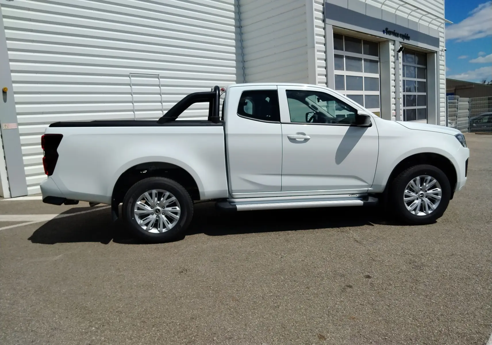 Isuzu D-MAX blanc en 3/4 avant droit, avec arceau et volet couvre benne roulant visible sur un parking extérieur.