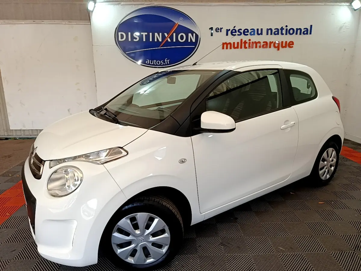 Citroën C1 blanc vue 3/4 avant droit, compacte avec phares ronds et rétroviseurs blancs, en intérieur showroom.