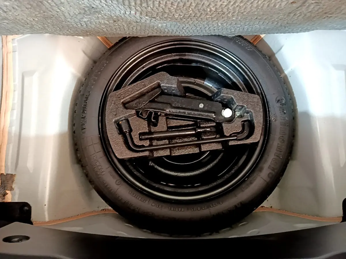 Vue plongeante sur la roue de secours noire et ses outils rangés dans le coffre d'une Citroën C1 blanche.