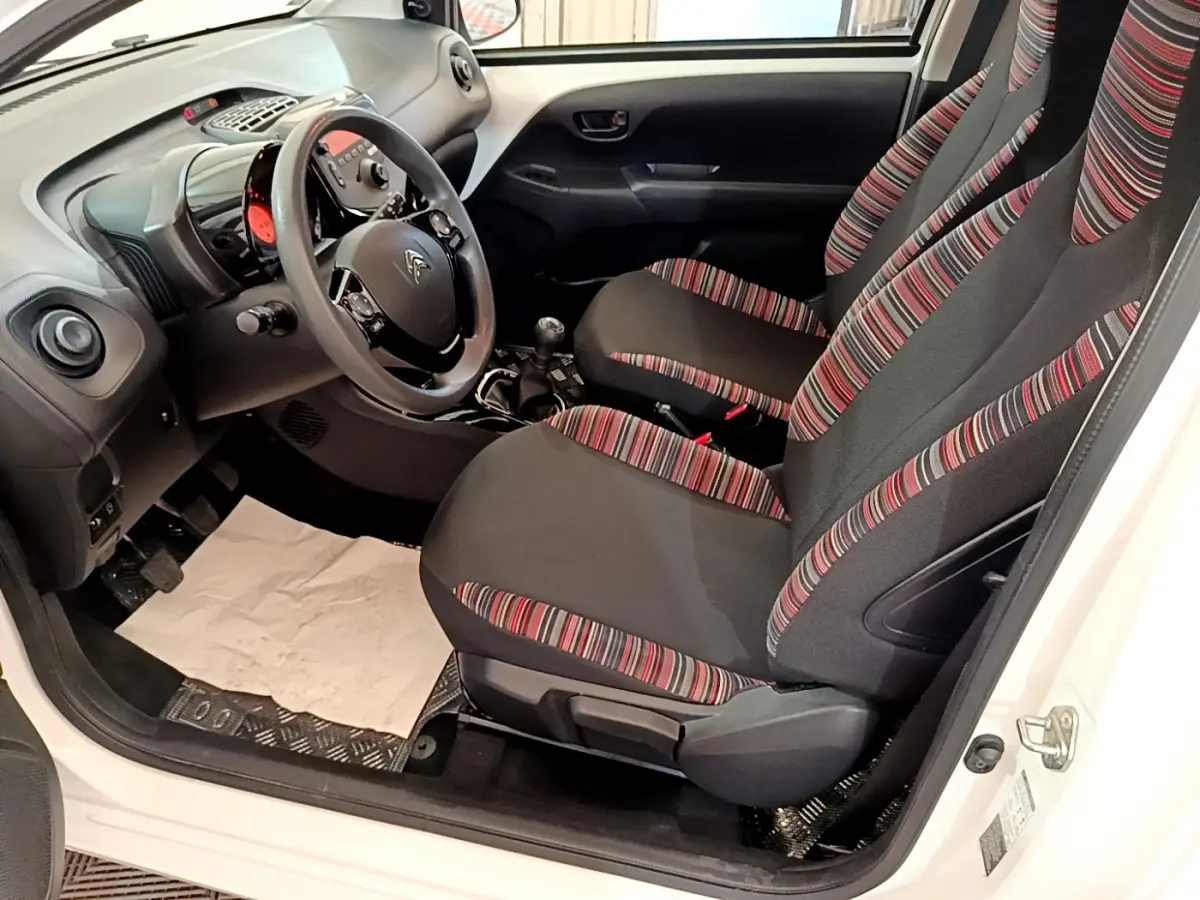 Vue intérieure côté conducteur de la Citroën C1 blanche 2020, sièges tissu noir à rayures rouges et volant multifonction.
