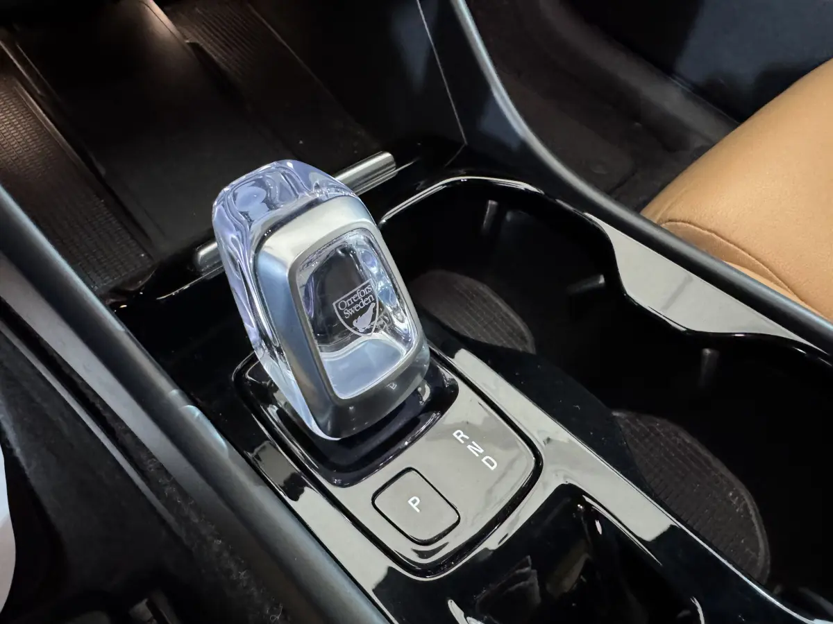 Gros plan sur la console centrale noire du Volvo XC40 Recharge T5 avec levier de vitesses en cristal Orrefors et siège cuir beige visible.