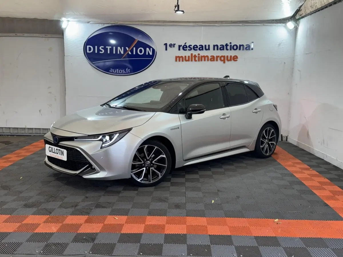 Toyota Corolla Hybride gris clair vue 3/4 avant droit en intérieur sur sol damier noir et orange.