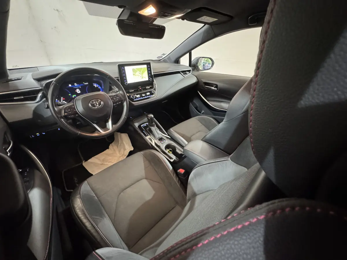 Vue intérieure avant droite de la Toyota Corolla hybride 2021, sièges cuir noir avec surpiqûres rouges et écran tactile central.