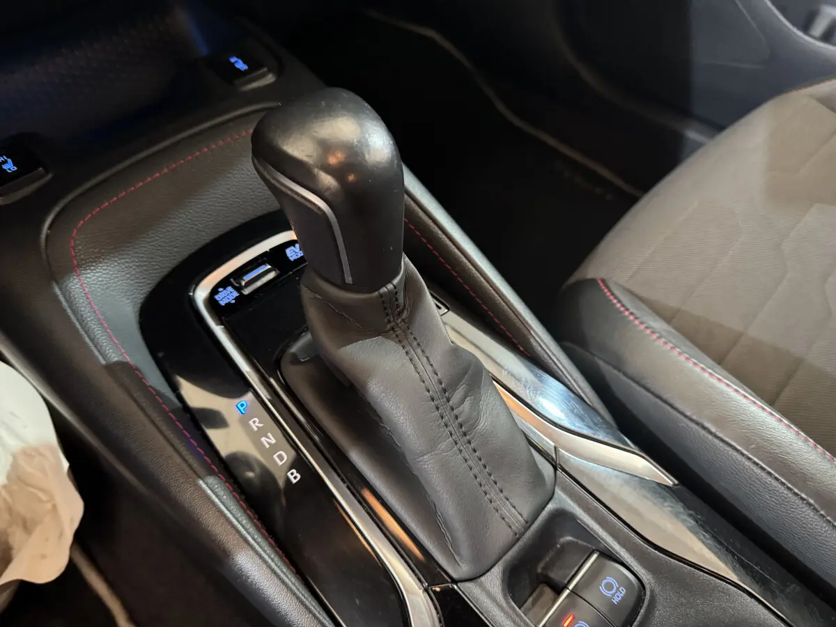 Gros plan sur la console centrale avec levier de vitesse automatique en cuir noir et surpiqûres rouges dans l’habitacle d’une Toyota Corolla gris clair.
