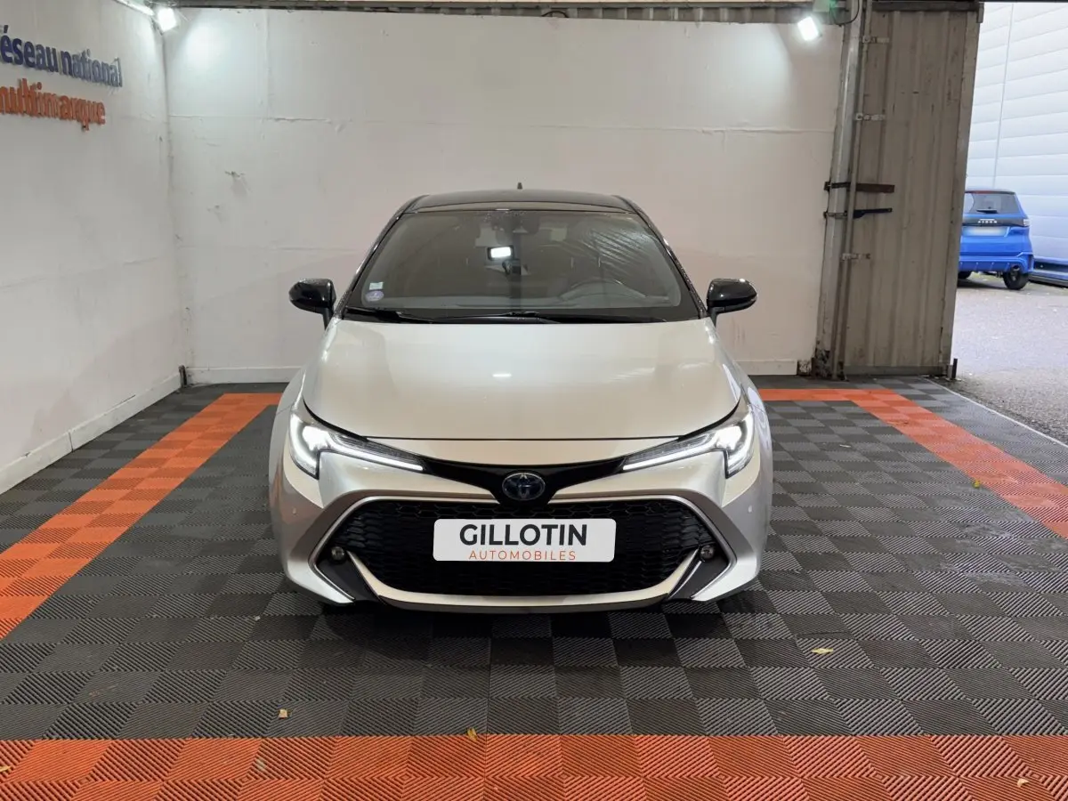 Vue avant frontale de la Toyota Corolla Hybride 122h Collection gris clair avec calandre noire et phares LED allumés.