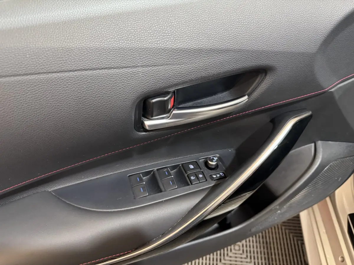 Gros plan sur la poignée intérieure et les commandes de porte côté conducteur d'une Toyota Corolla gris clair, avec surpiqûres rouges.
