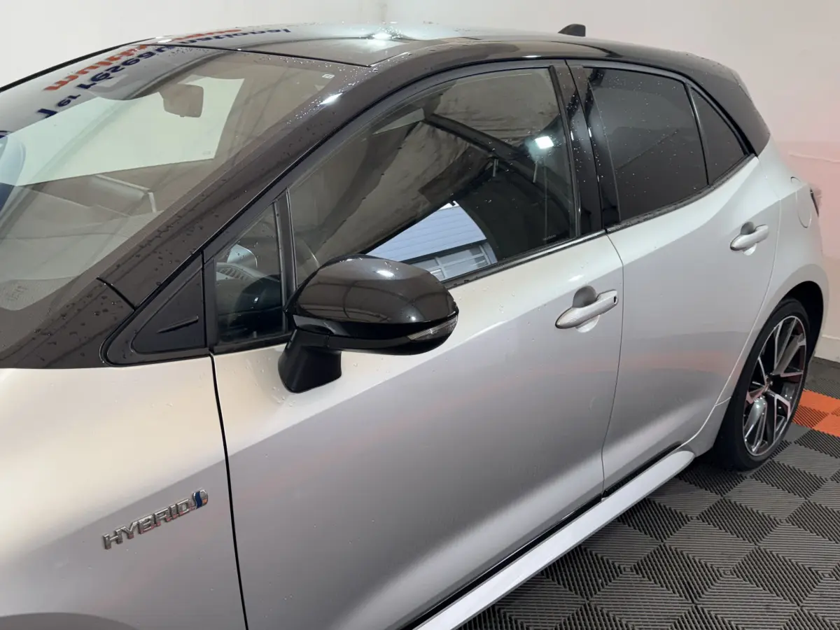 Vue côté gauche d’une Toyota Corolla Hybride gris clair avec toit et rétroviseur noir brillant, badge Hybrid visible.