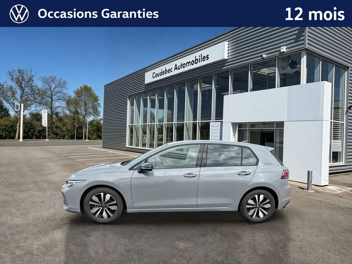 Profil côté gauche d'une Volkswagen Golf 1.5 eTSI 150 DSG7 grise, garée devant un bâtiment moderne de concession.