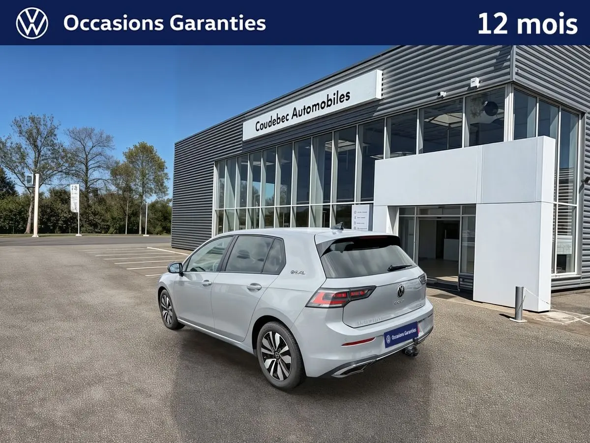 Vue 3/4 arrière droite d'une Volkswagen Golf grise stationnée devant un bâtiment moderne avec attelage escamotable visible.