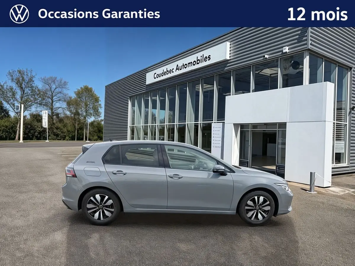 Vue latérale droite d'une Volkswagen Golf grise stationnée devant un bâtiment moderne en verre.