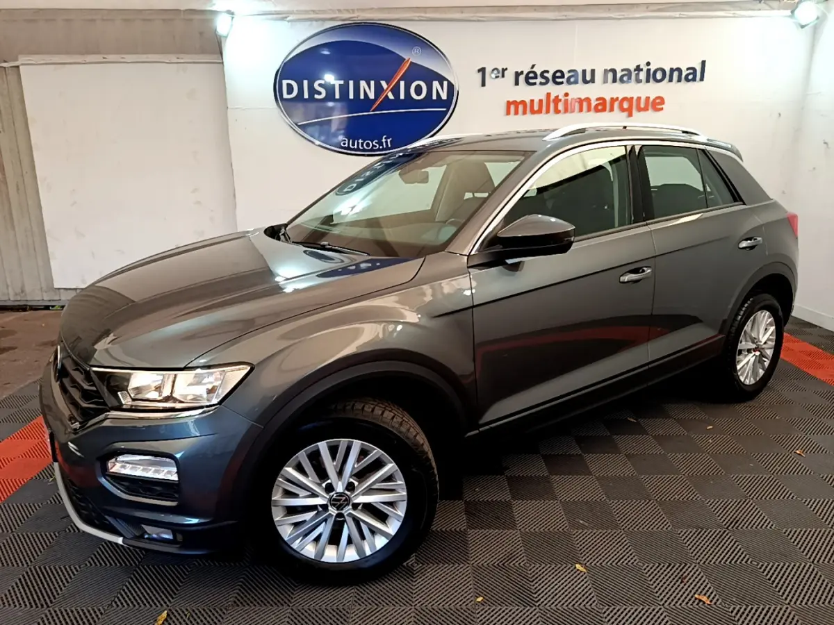 Volkswagen T-Roc gris foncé vue 3/4 avant droit, avec jantes alliage et barres de toit argentées visibles.