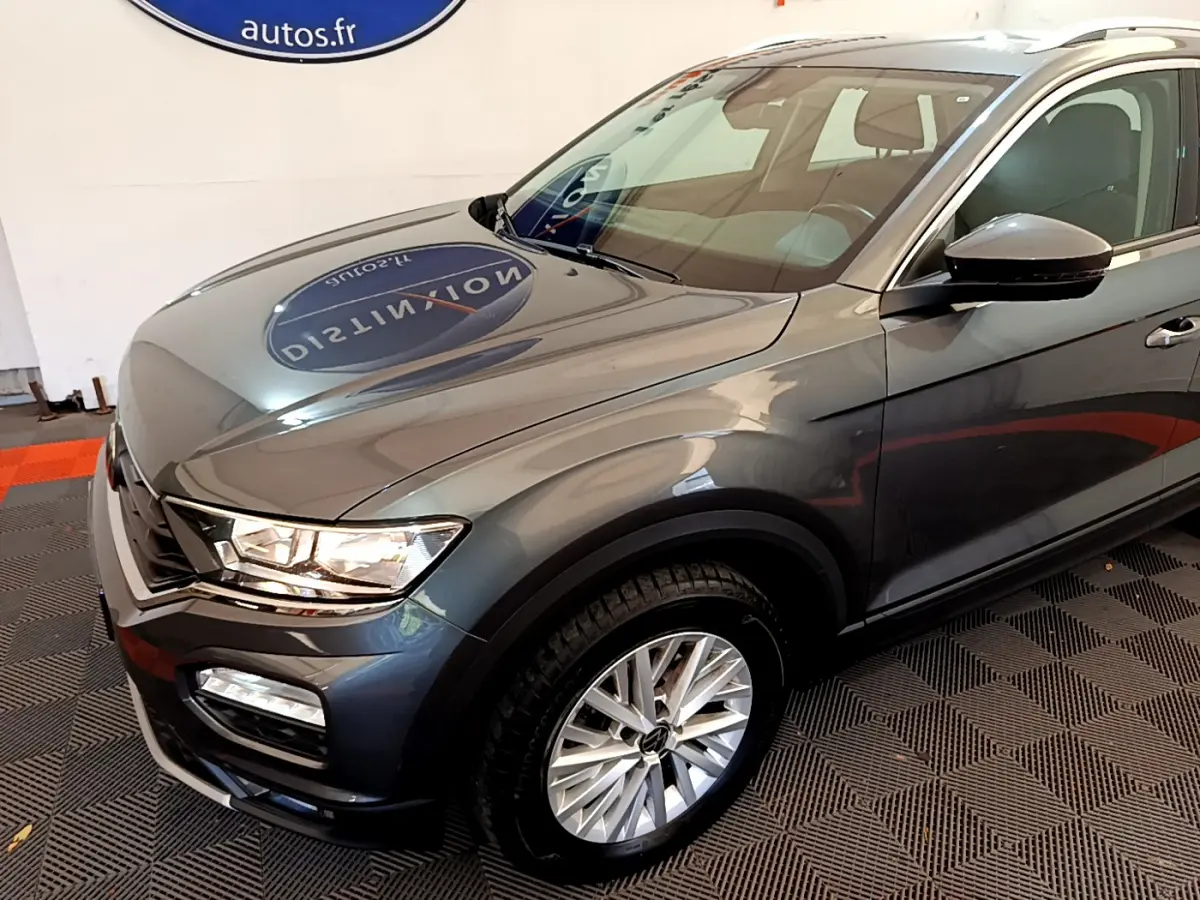 Vue 3/4 avant droit d'un Volkswagen T-Roc gris foncé avec jantes alliage et phares LED visibles en intérieur showroom.