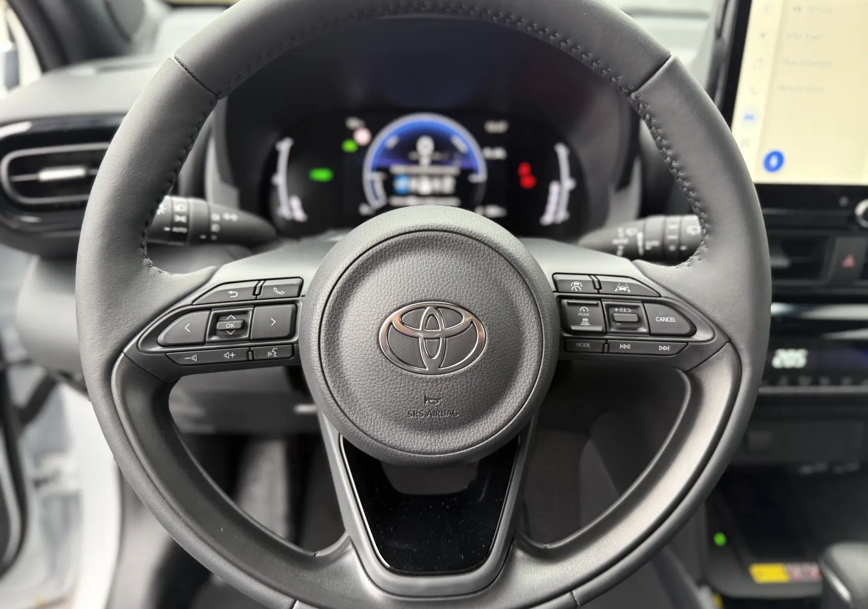 Vue rapprochée du volant noir de la Toyota Yaris Cross 130 CH Design 2025 avec commandes intégrées et tableau de bord numérique flou.