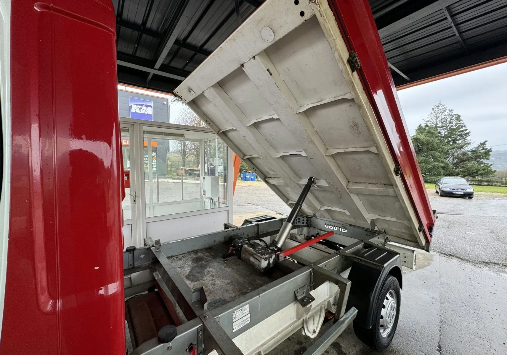 Vue arrière droite d'un Citroën Jumper benne rouge avec benne relevée montrant le châssis et le vérin hydraulique.