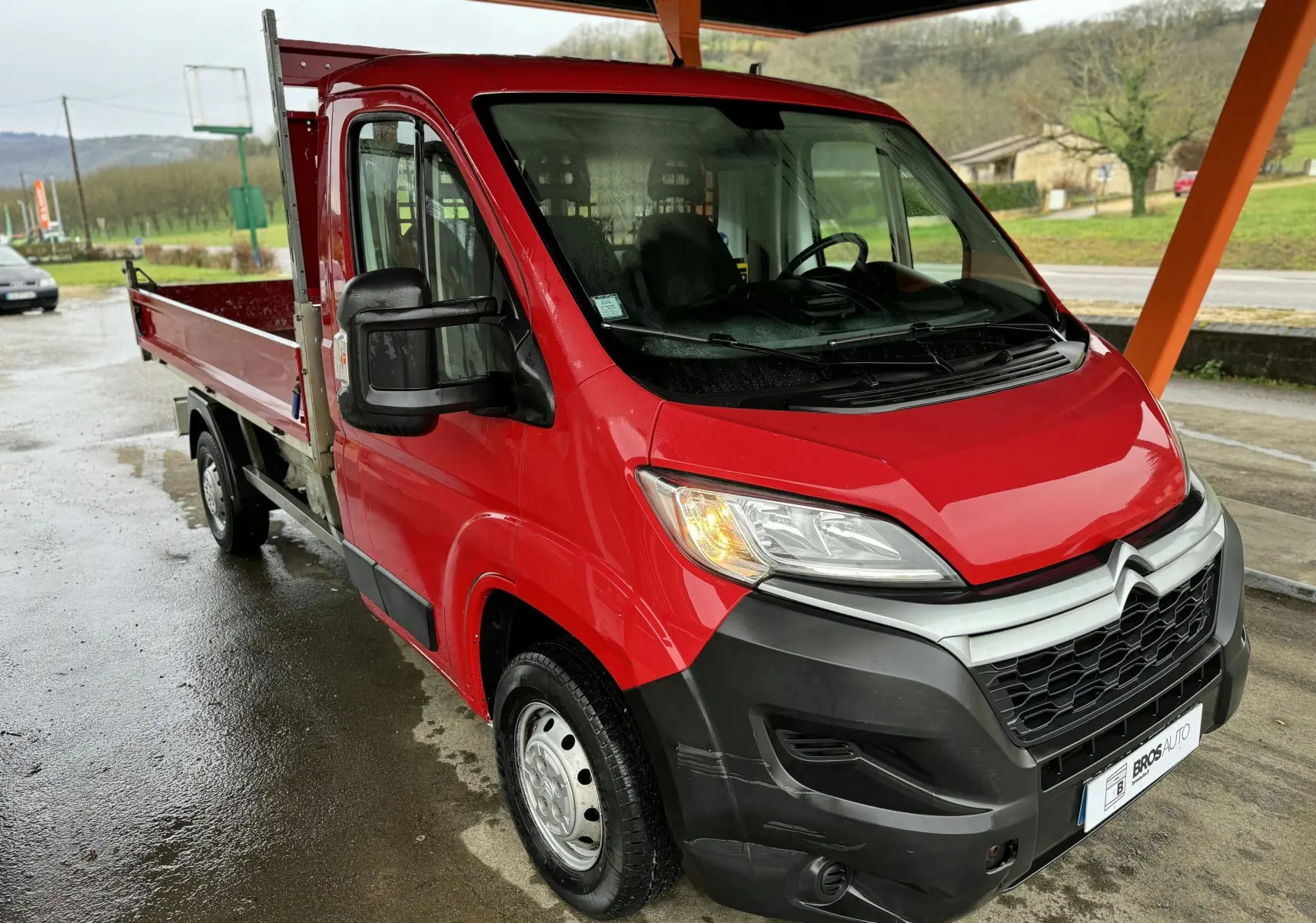 Vue 3/4 avant gauche d'un Citroën Jumper benne rouge avec ridelles latérales sous un abri.