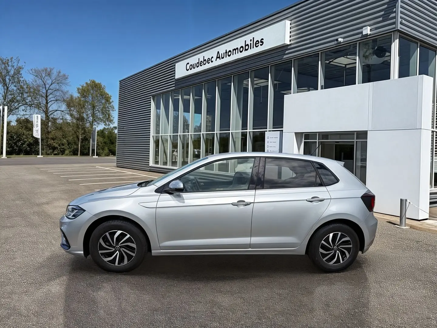 Profil droit d'une Volkswagen Polo grise 2025 stationnée devant un bâtiment moderne de concession automobile.