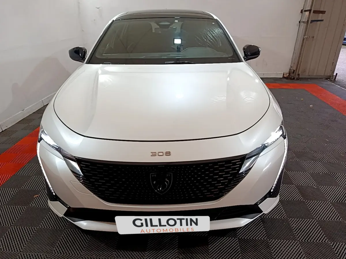 Vue frontale d'une Peugeot 308 PHEV 180 E-EAT8 GT blanche avec calandre noire et phares LED allumés dans un garage.