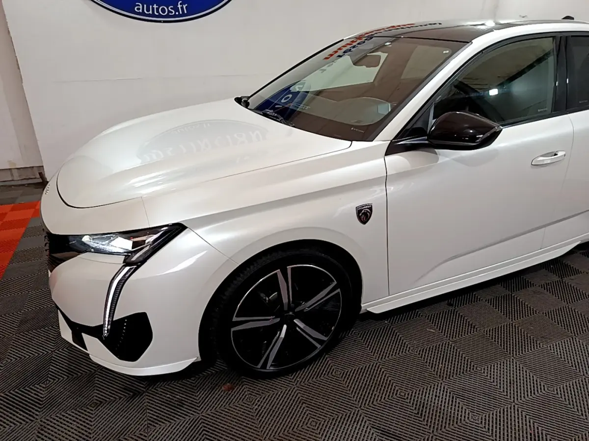 Vue 3/4 avant droit d'une Peugeot 308 PHEV 180 GT blanche avec jantes noires et logo GT sur l'aile avant.