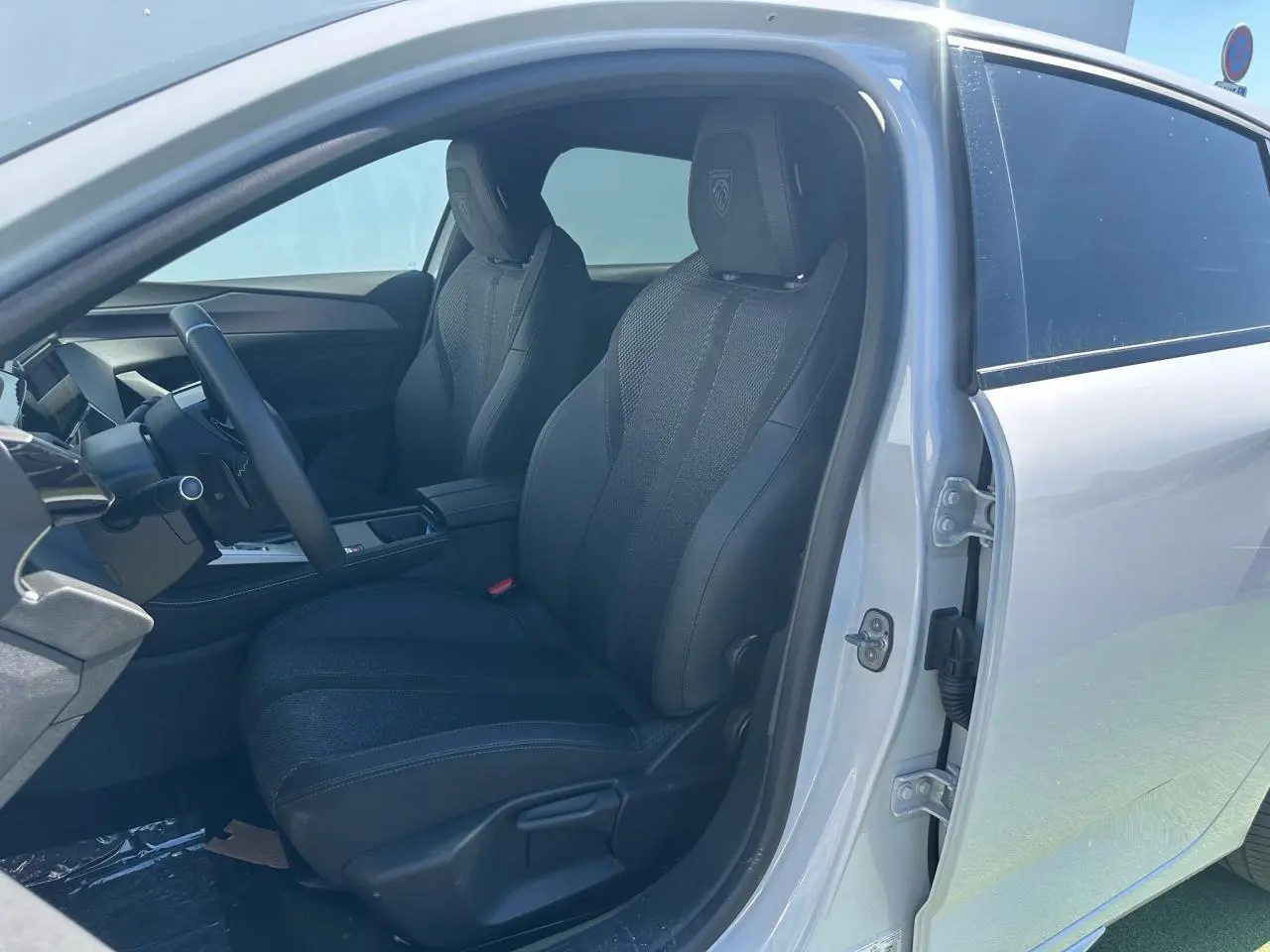 Vue intérieure côté conducteur de la Peugeot 308 blanche Okenite, sièges noirs avec logo Peugeot brodé sur appuie-tête.