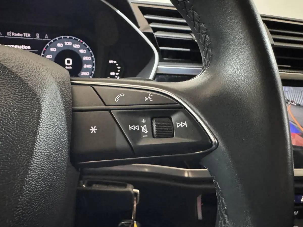 Gros plan sur le volant noir de l'Audi Q3 2022, avec commandes audio et affichage numérique partiel en arrière-plan.