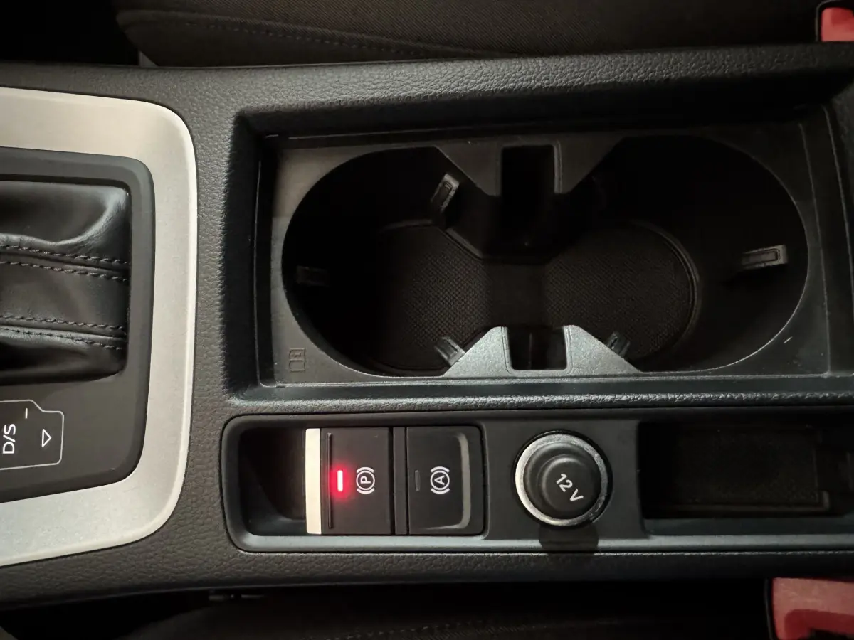 Gros plan sur la console centrale noire de l'Audi Q3 2022, avec porte-gobelets, bouton frein à main électrique et prise 12V.