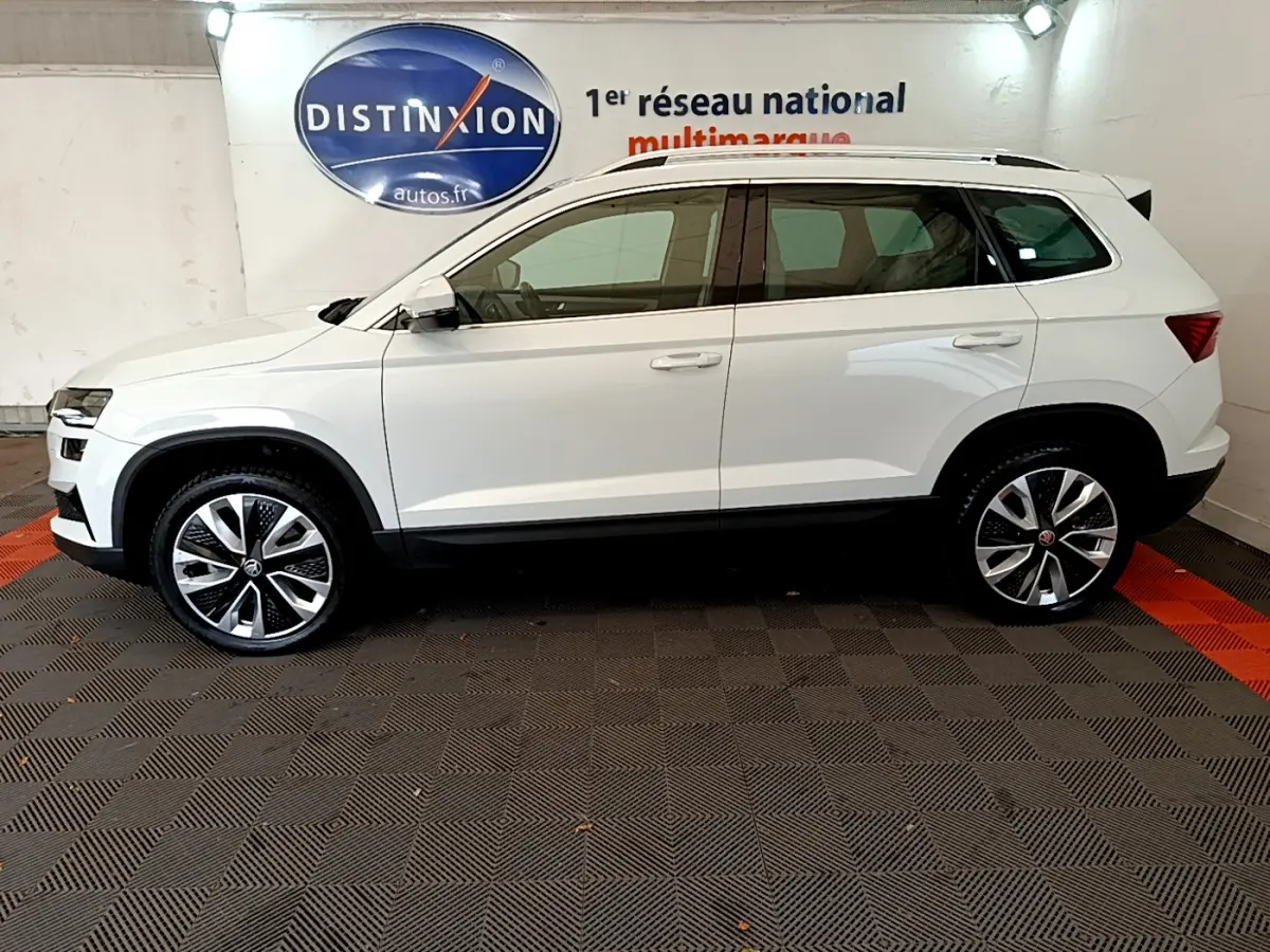Vue latérale droite d'un Skoda Karoq blanc 2023 avec jantes alliage et rails de toit chromés en intérieur showroom.