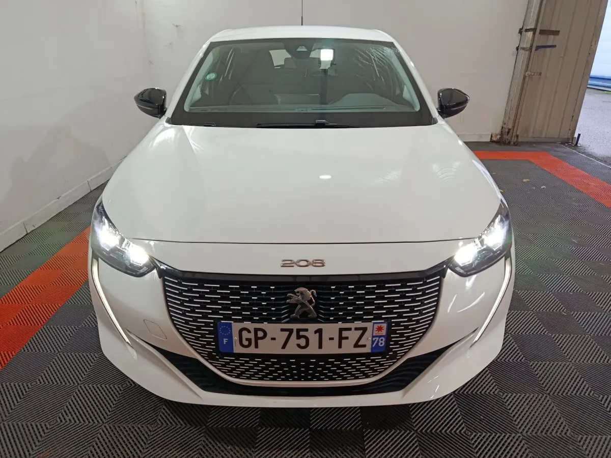 Vue frontale d'une Peugeot 208 électrique blanche avec phares LED allumés et calandre noire distinctive.