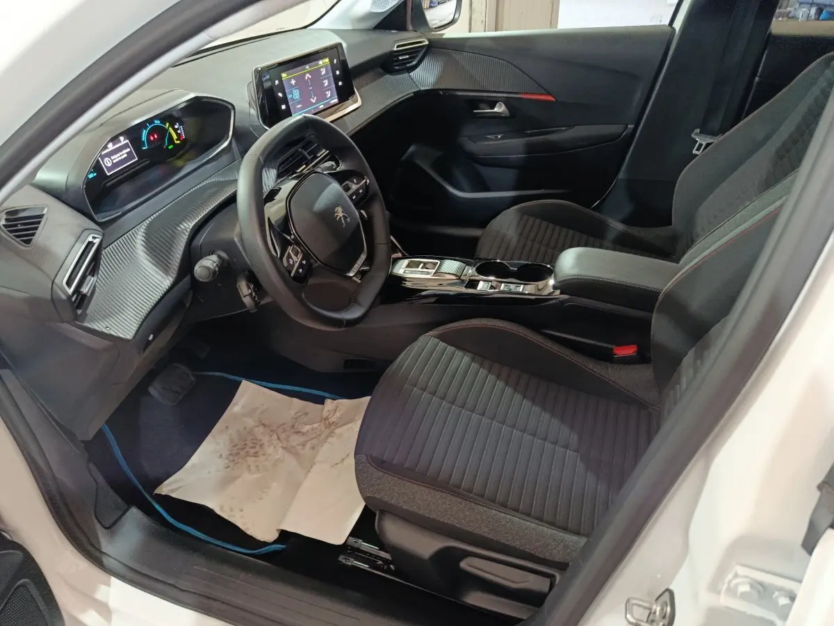 Intérieur de la Peugeot 208 électrique blanche, vue côté conducteur, avec tableau de bord digital et sièges noirs à surpiqûres rouges.
