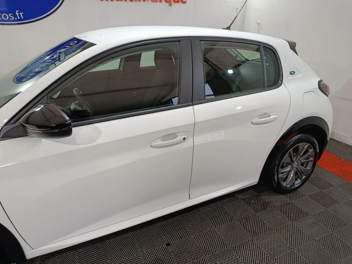 Profil côté gauche d'une Peugeot 208 électrique blanche 2023, avec rétroviseur noir et jantes alliage visibles.