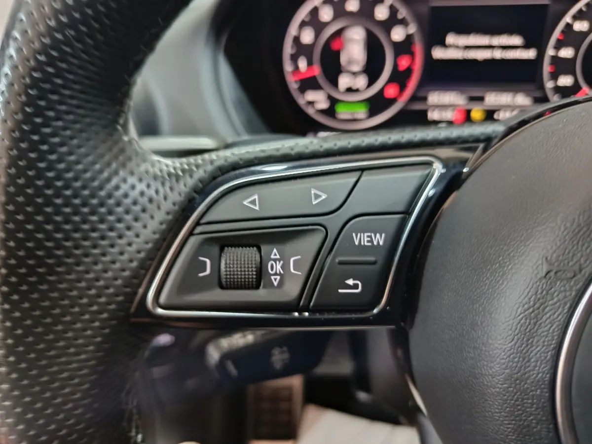 Gros plan sur les commandes gauche du volant cuir perforé d'un Audi Q2 noir, avec tableau de bord numérique en arrière-plan.