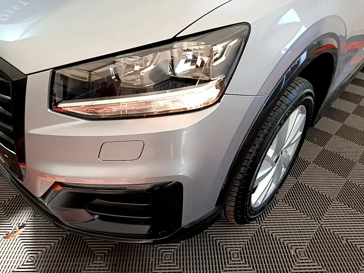 Gros plan sur l'avant droit gris clair de l'Audi Q2 35 TDI 150 Design, mettant en valeur le phare et la roue.