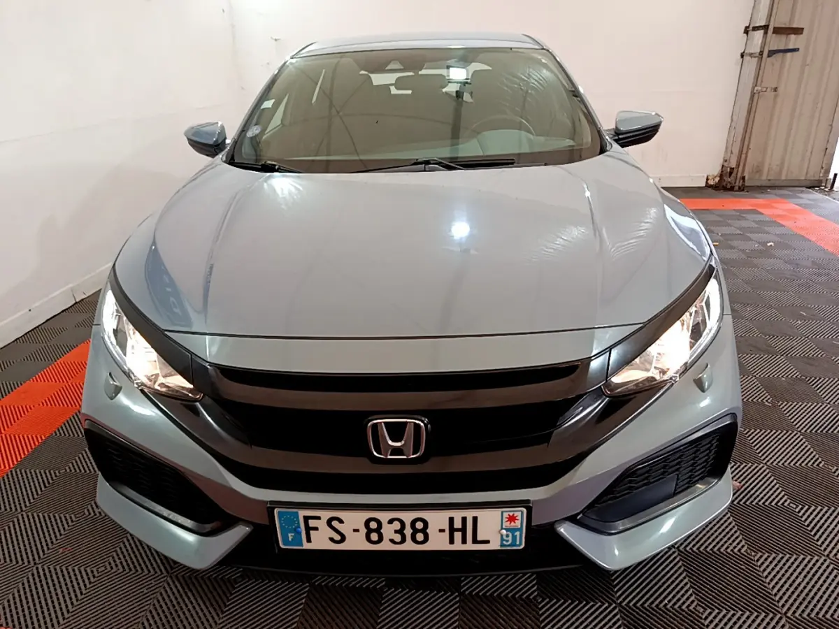 Vue frontale d'une Honda Civic gris foncé avec phares allumés et calandre noire brillante dans un garage.