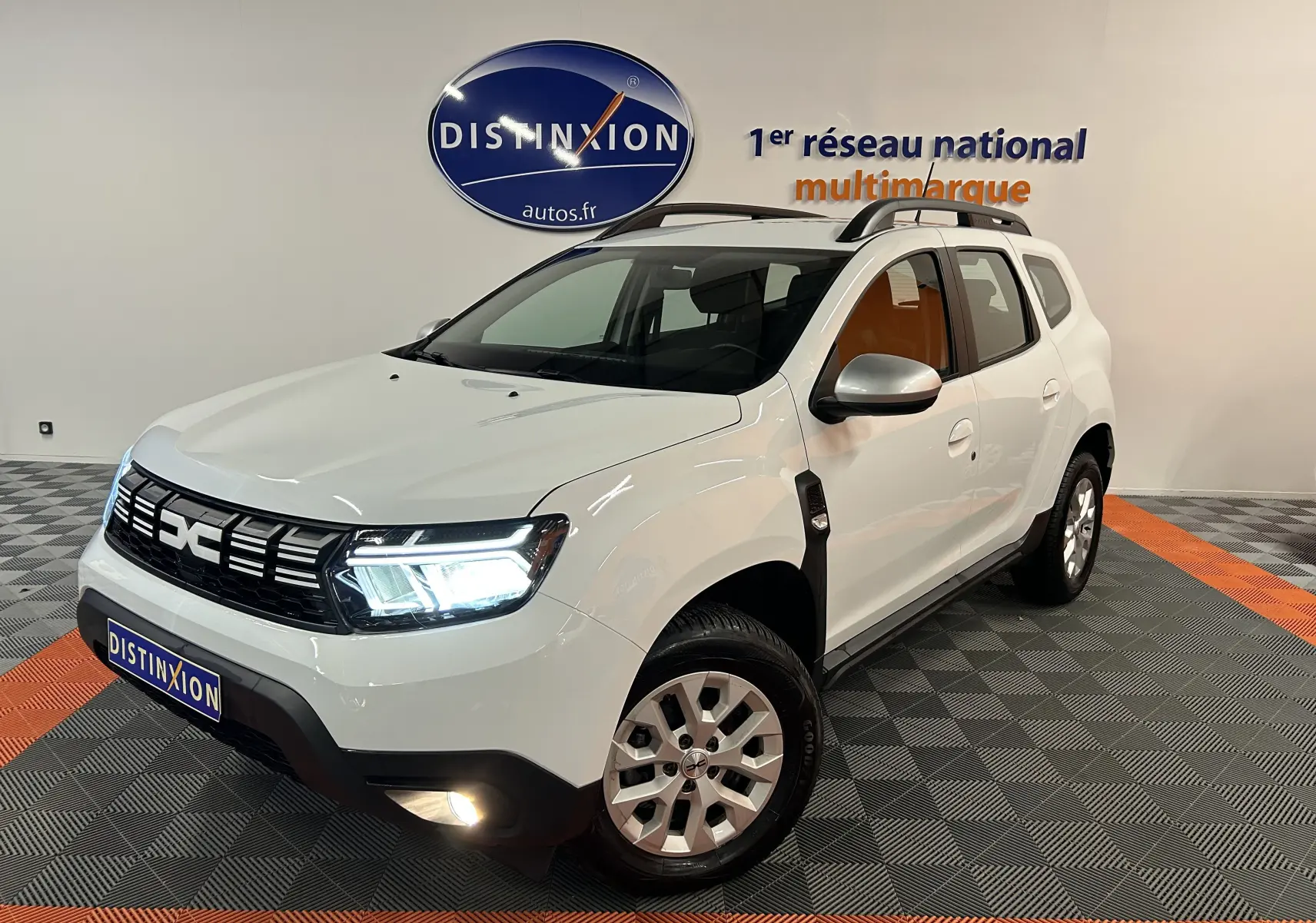 Vue 3/4 avant droite d'un Dacia Duster blanc 2024 avec feux LED allumés et barres de toit noires.